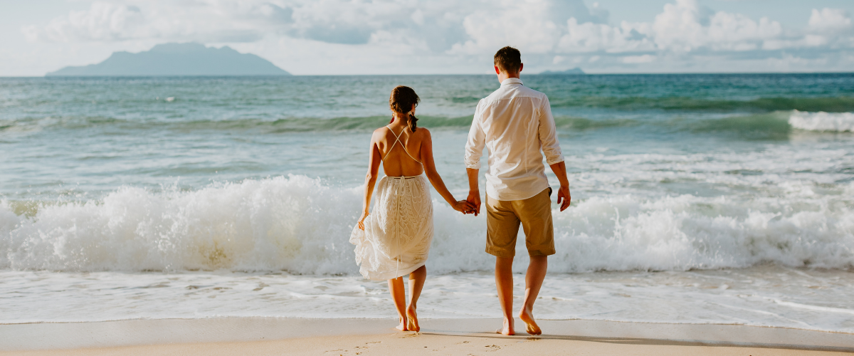 Bride and groom holding hands walking along the beach toward the ocean
