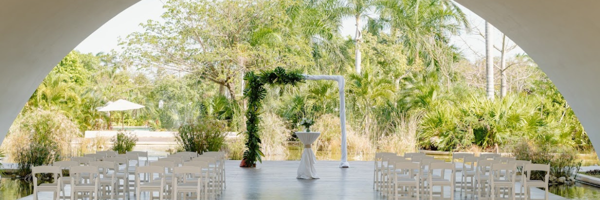 Destination wedding set up under a covering with white chairs and a simple arch with green leafy arrangement around half of it and palm trees in the background