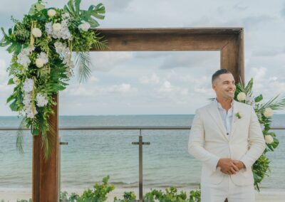 groom wearing a white suit and smiling in front of the wedding arch setup for his ceremony, destination wedding at the beach on a cloudy day.