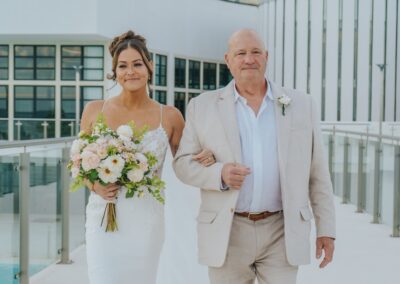 bride smiling walking next to her proud father down the aisle at her destination wedding. White hotel seen in the background.