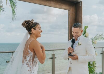 groom looking at his phone reading his wedding vows to his bride. Both are smiling and laughing. Square wedding arch and beach scene in the background.