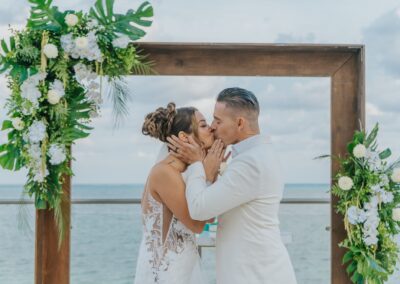 couples first wedding kiss, their wedding arch and green and white florals as the backdrop by the beach.