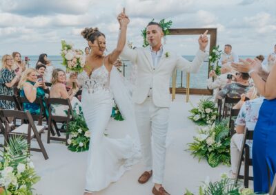 bride and groom smiling holding hands with their arms up after getting married. Seated guests clapping and taking pictures.
