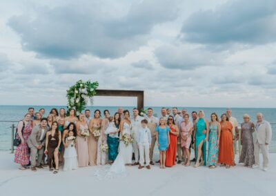 group photo at a destination wedding. Guests are wearing different colored outfits, beach and cloud scene.