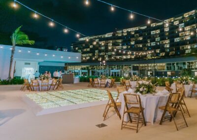 set up at a hotel resort for a destination wedding reception at night. Tables with white table cloths and wooden chairs set up all around.