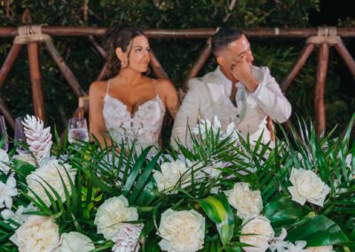 bride and groom sitting at their wedding reception listening to a toast, huge white and green florals can be seen in the foreground.