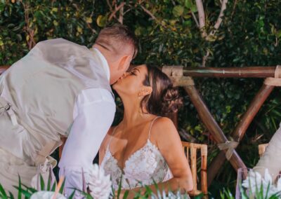 couple at wedding reception, bride kissing groom on the cheek. Tree and leaves seen in the background.