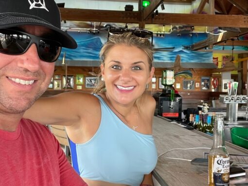 Heather and David D'Amico from D'Amico Travel. They are looking at the camera and smiling, sitting at a bar while on vacation.