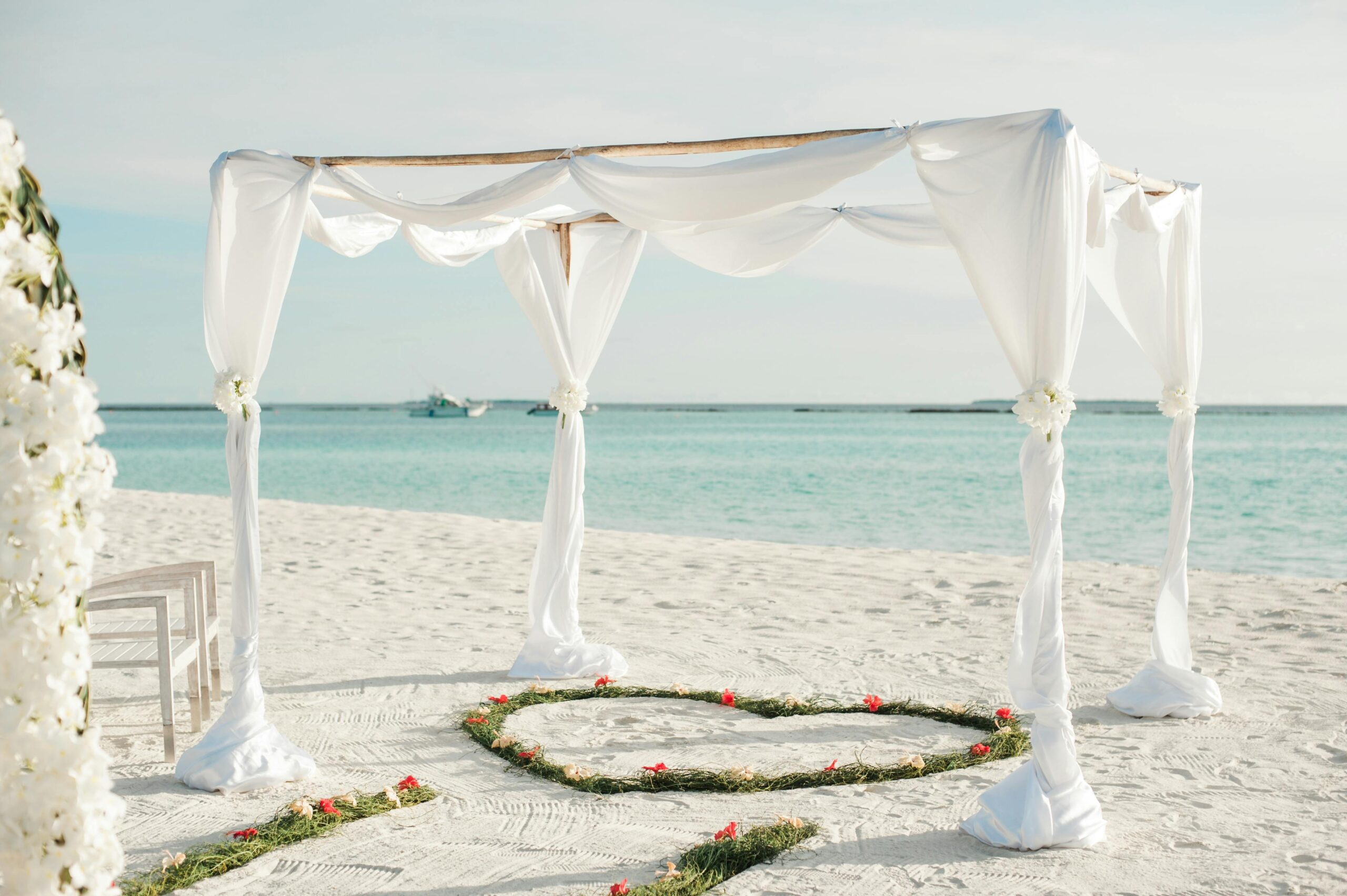 destination-wedding-asadphoto-pexels beautiful white wedding arch set up at a white sand beach for a destination wedding. A heart shape is made in the sand with leaves and flowers. Turquoise waters in the background.