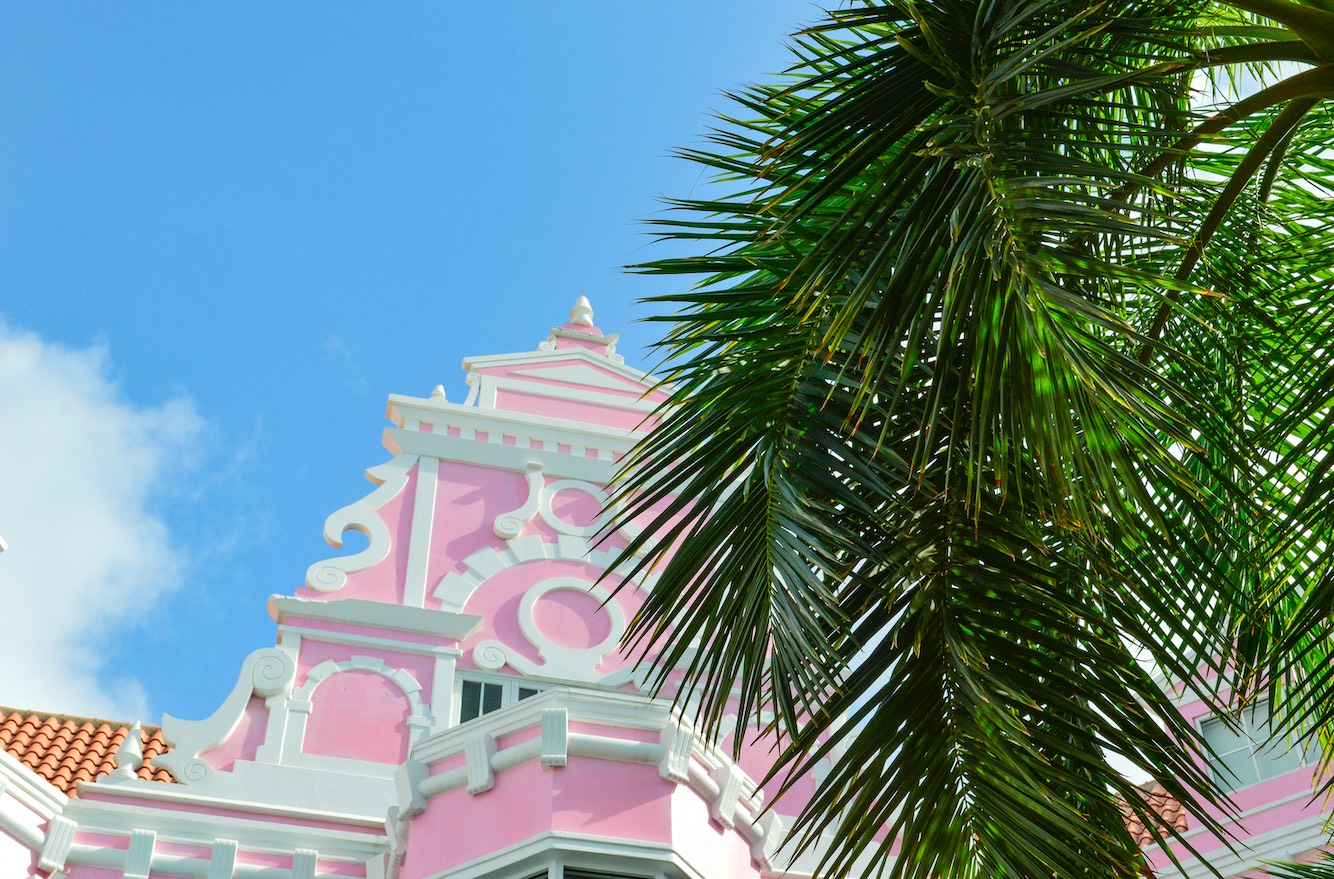 Aruba pink pastel pink pastel building behind a palm tree in Aruba. Blue Caribbean skies.