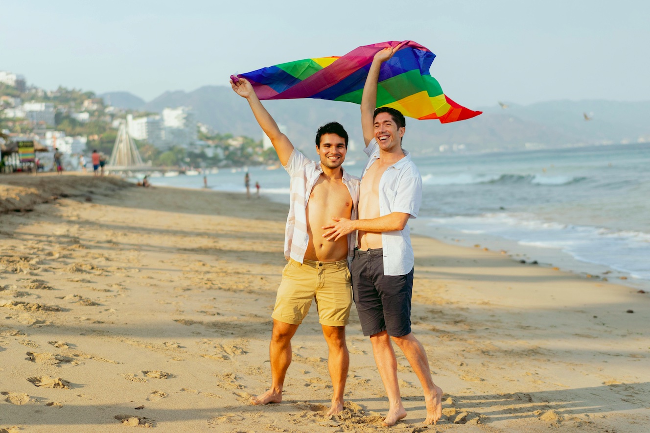 Puerto Vallarta pride flag two queer men holding up the Pride flag and smiling with happiness at a beach in Puerto Vallarta, Mexico, a very welcoming place for gay destination weddings.